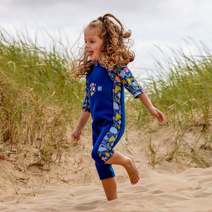 Lifestyle image of child wearing a one piece UV sun and sea wetsuit for toddlers in navy blue with yellow trims. Flowers and insects themed print on sleeves, side panels and neck. 