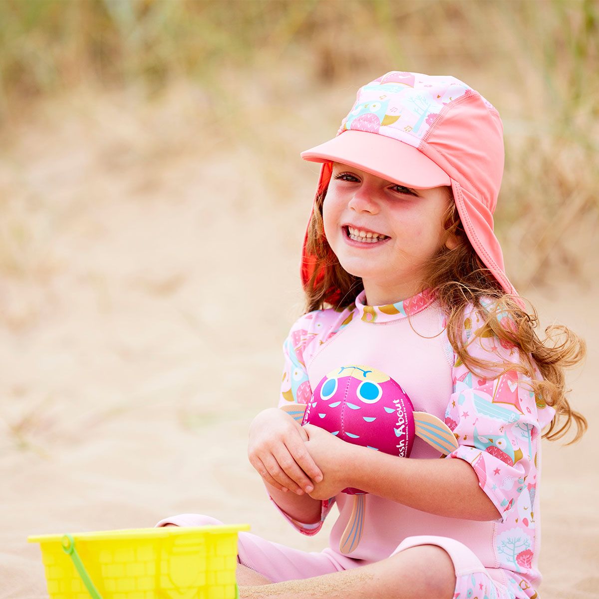 Lifestyle image of toddler wearing a legionnaire style sun hat in the beach. The hat is reddish pink and baby pink, with the owl and the pussycat themed print panel. She's also wearing matching sun and sea wetsuit. Front.
