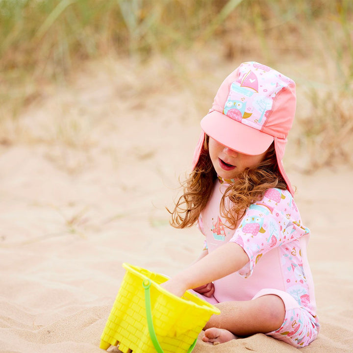 Lifestyle image of toddler wearing a legionnaire style sun hat in the beach. The hat is reddish pink and baby pink, with the owl and the pussycat themed print panel. She's also wearing matching sun and sea wetsuit.