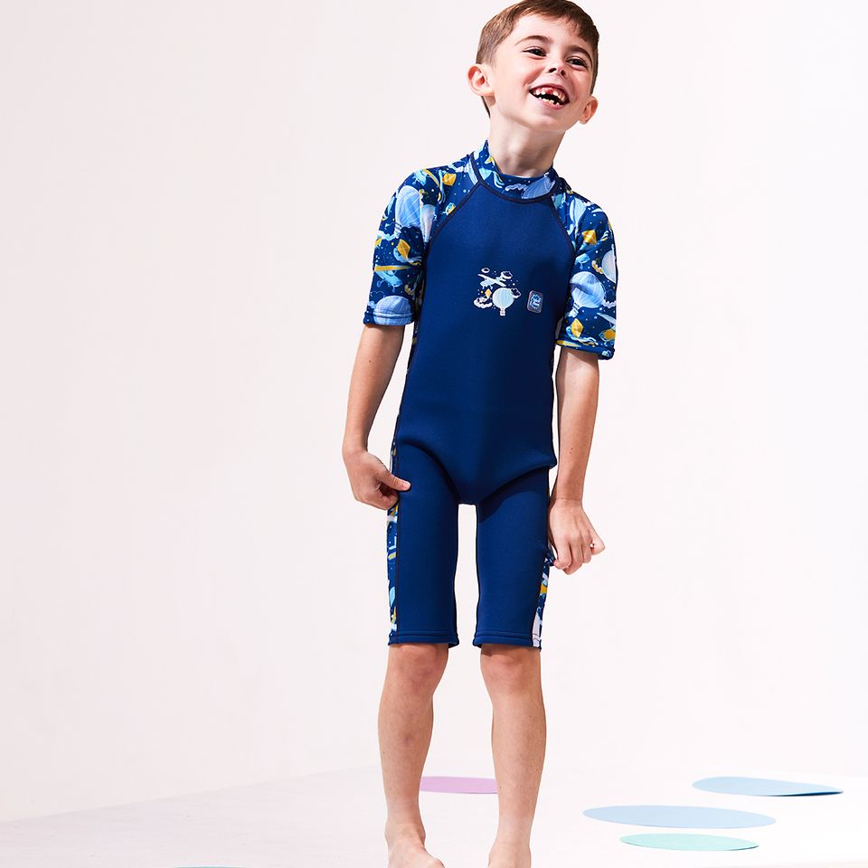 Lifestyle image of child wearing a wne piece UV sun and sea wetsuit  in navy blue. Sky themed print including airplanes, air hot balloons, clouds and kites on sleeves, side panels and neck. Front.