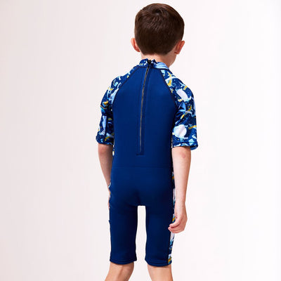 Lifestyle image of child wearing a wne piece UV sun and sea wetsuit  in navy blue. Sky themed print including airplanes, air hot balloons, clouds and kites on sleeves, side panels and neck. Back.