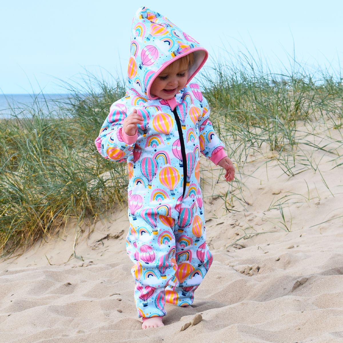 Lifestyle image of toddler wearing a waterproof fleece-lined onesie with hood in baby blue and hot air balloons themed print, including rainbows and clouds. Pink trims. 