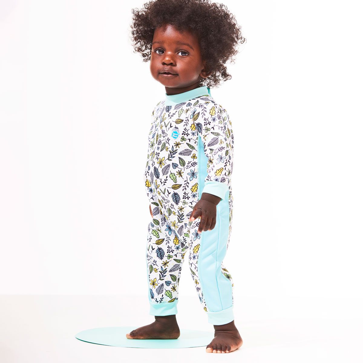 Lifestyle image of toddler wearing a fleece-lined baby wetsuit in white with light blue trims and leaves themed print. 