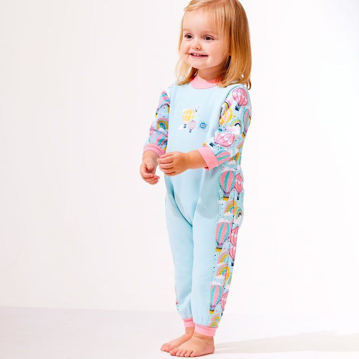 Lifestyle image of toddler wearing a fleece-lined baby wetsuit in baby blue with pink trims and hot air balloons themed print, including rainbows and clouds. 