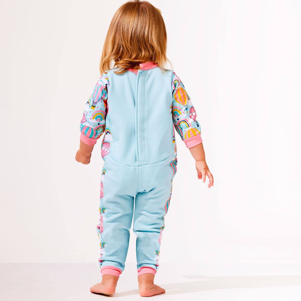Lifestyle image of toddler wearing a fleece-lined baby wetsuit in baby blue with pink trims and hot air balloons themed print, including rainbows and clouds. 