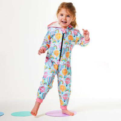 Lifestyle image of toddler wearing a waterproof fleece-lined onesie with hood in baby blue and hot air balloons themed print, including rainbows and clouds. Pink trims. 